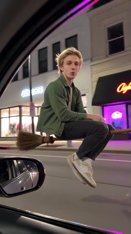 Still from a generated clip: a young man in a green jacket by a parked car on a lit night street, shop windows behind him