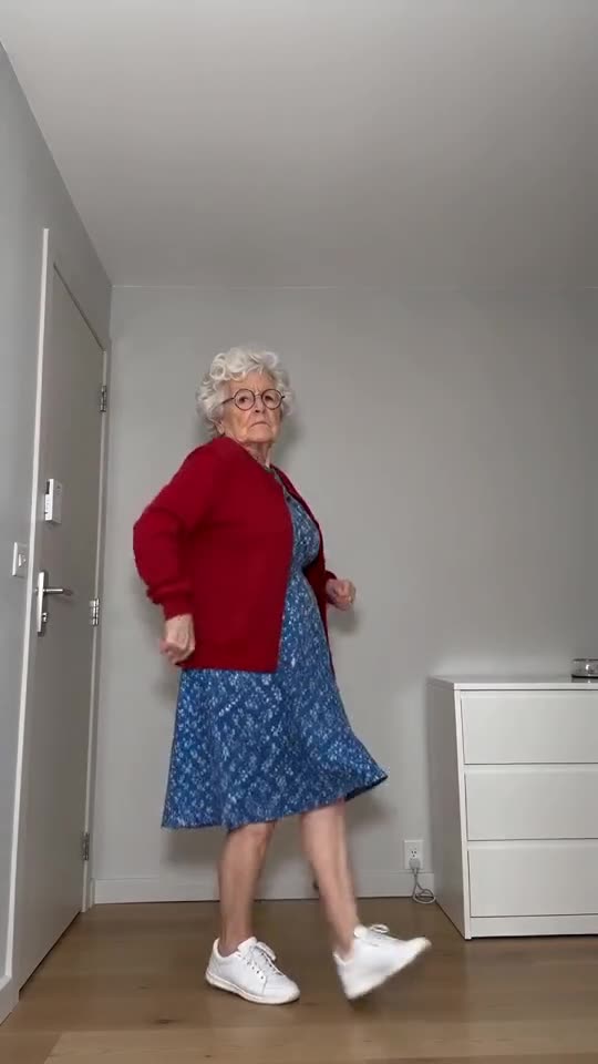 Still from the sample: a made-up grandmother with white curls, round glasses, a red cardigan and a floral dress, captured mid-hop and expressionless beside a white door in a bare room